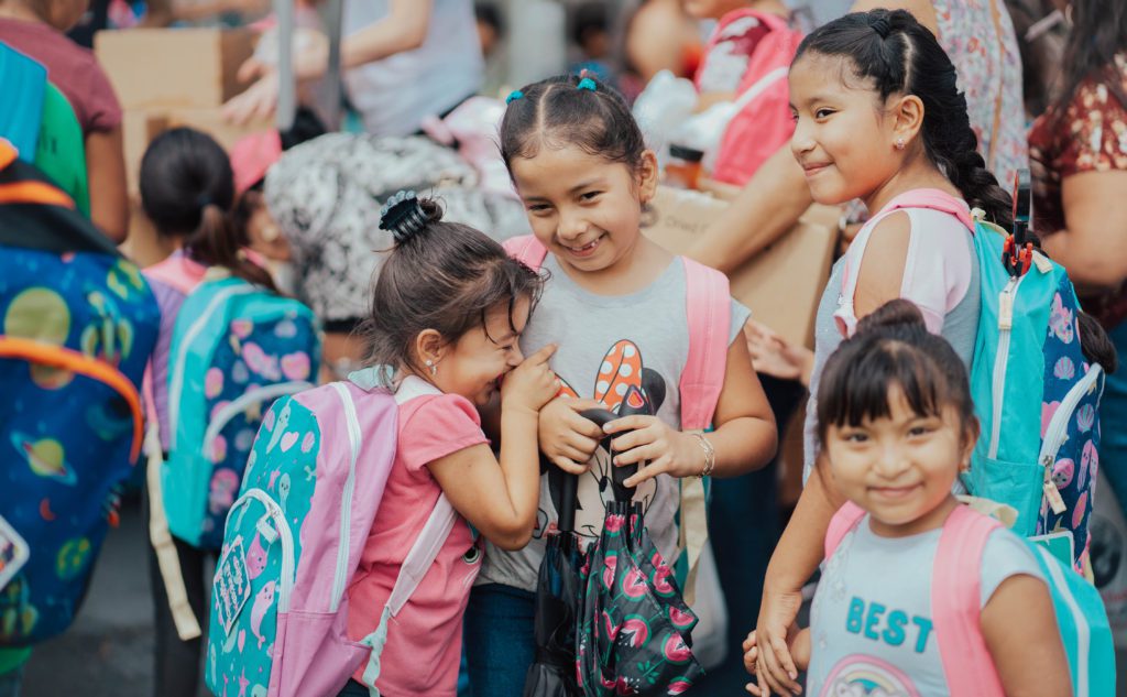 Four young girls smiling with backpacks on in crowd